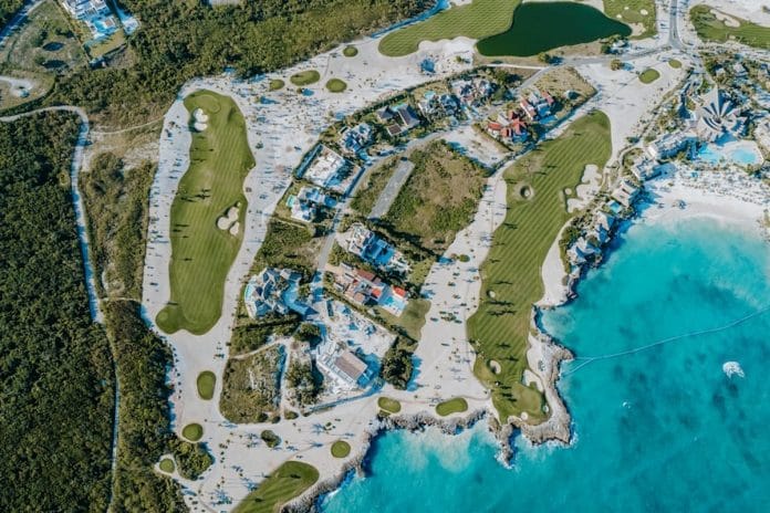 A par-three island green golf hole surrounded by water, with a small bridge leading out to the green and a sand bunker visible at its edge.
