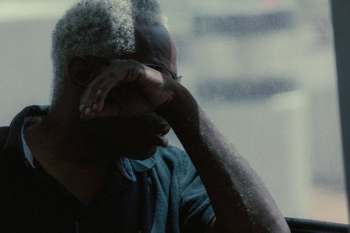 An older man sits close to a television showing a college basketball broadcast, with a notepad on his lap.