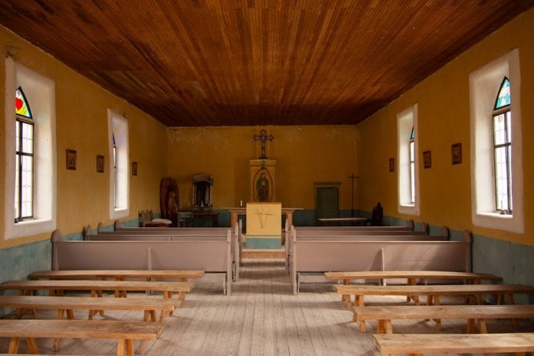 The interior of a small rural Catholic church with wooden pews and modest stained glass, soft morning light coming through the windows.