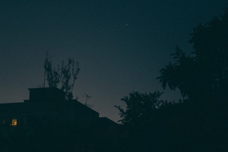 A quiet residential cul-de-sac at dusk with a single distant burst of fireworks above the rooftops