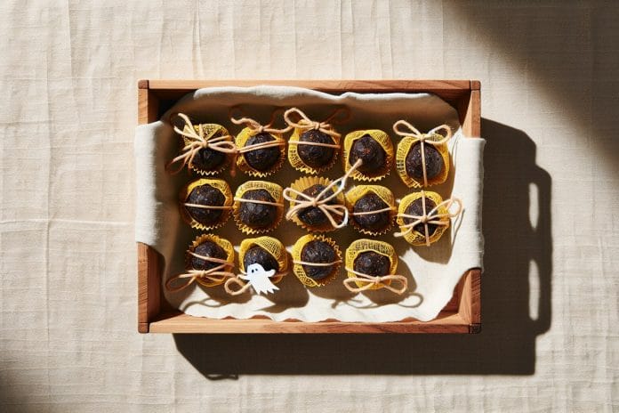 Beeswax-wrapped date balls with ghost stickers A tray of cacao-coated date balls wrapped in golden beeswax cloth tied with twine, each topped with a small paper ghost sticker.