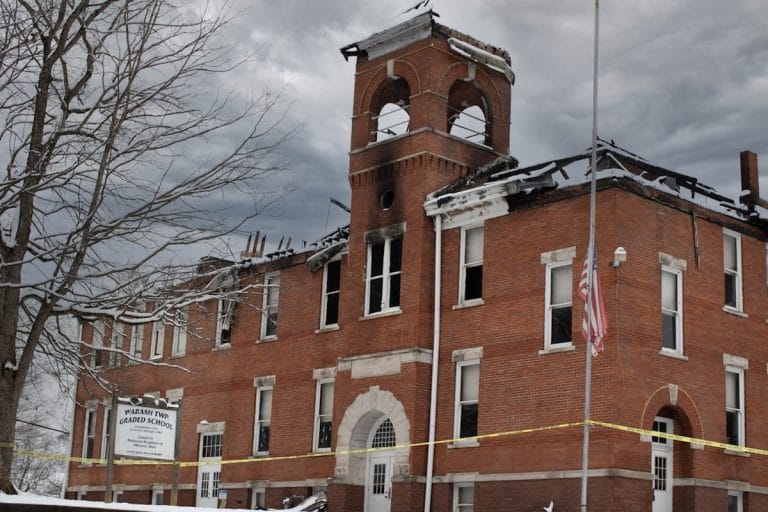 An older brick elementary school building on a quiet small-town street, flag pole out front, empty bus loop visible to one side.