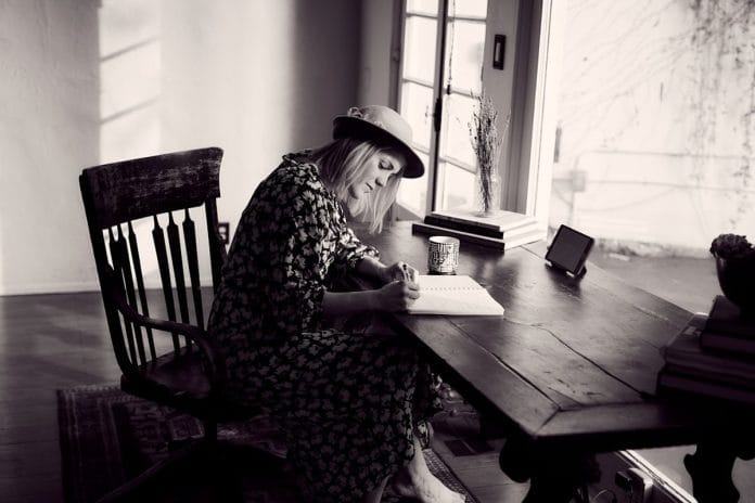 An older woman stands in a tidy kitchen holding a beige landline telephone next to a doily and a wall calendar.