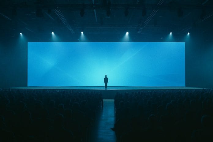 A lone executive stands center stage at a CES keynote, dwarfed by a vast glowing blue LED wall.
