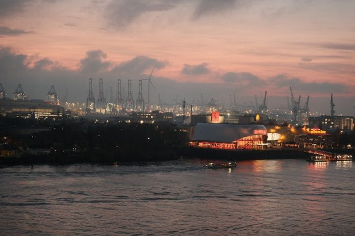 Stacked shipping containers at a commercial port at dawn, with gantry cranes silhouetted against a pale sky.