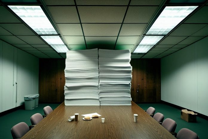 A towering stack of unbound printer paper on a government conference table under harsh fluorescent light, with empty coffee cups and a half-eaten sandwich beside it.
